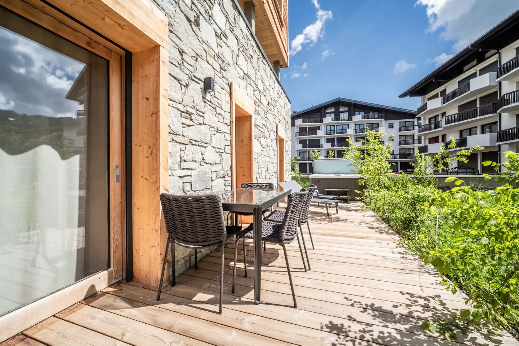 A wooden deck with outdoor dining furniture sits beside a stone-faced building and green plants.