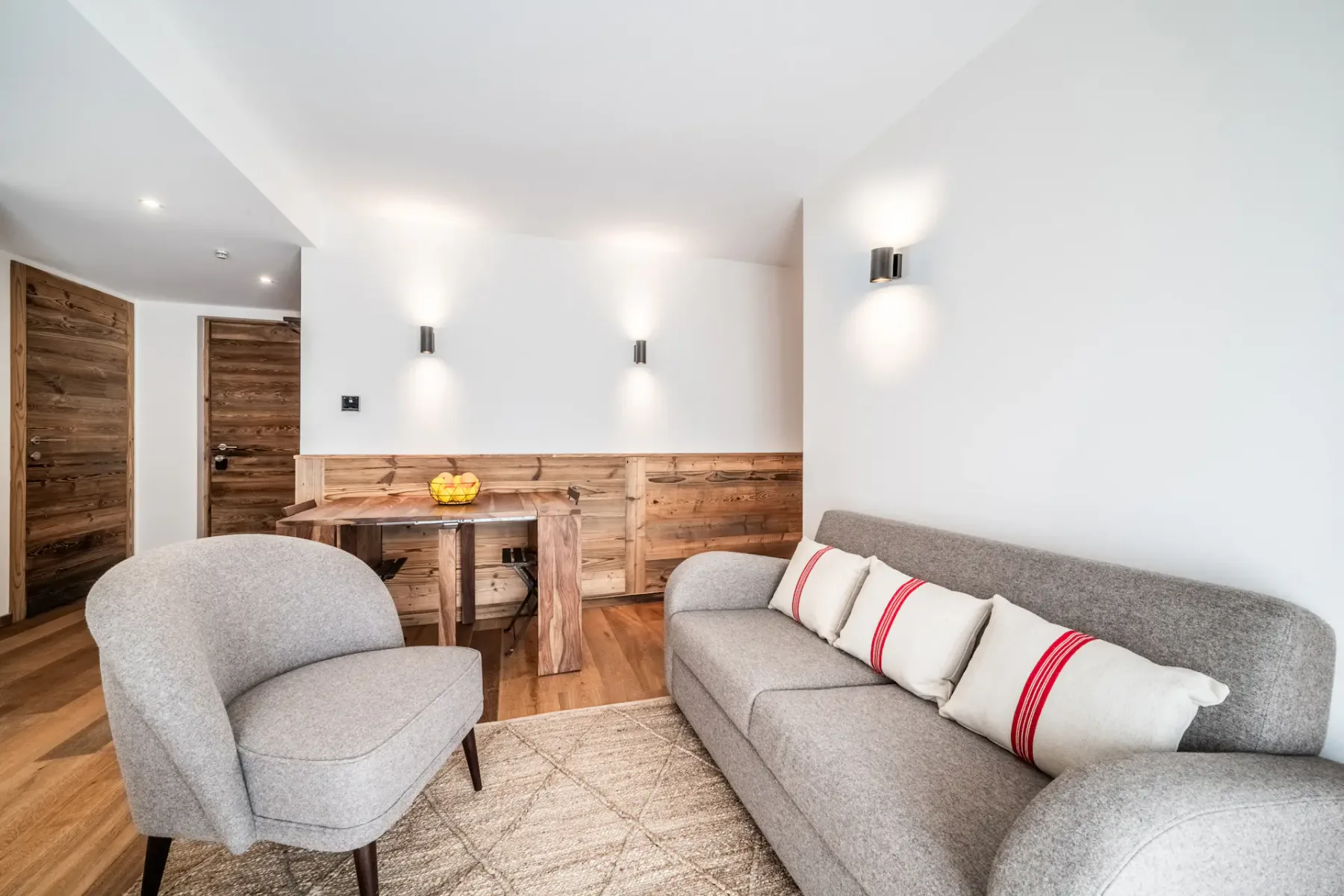 A bright living and dining area with gray seating, a wooden table, and a kitchenette.