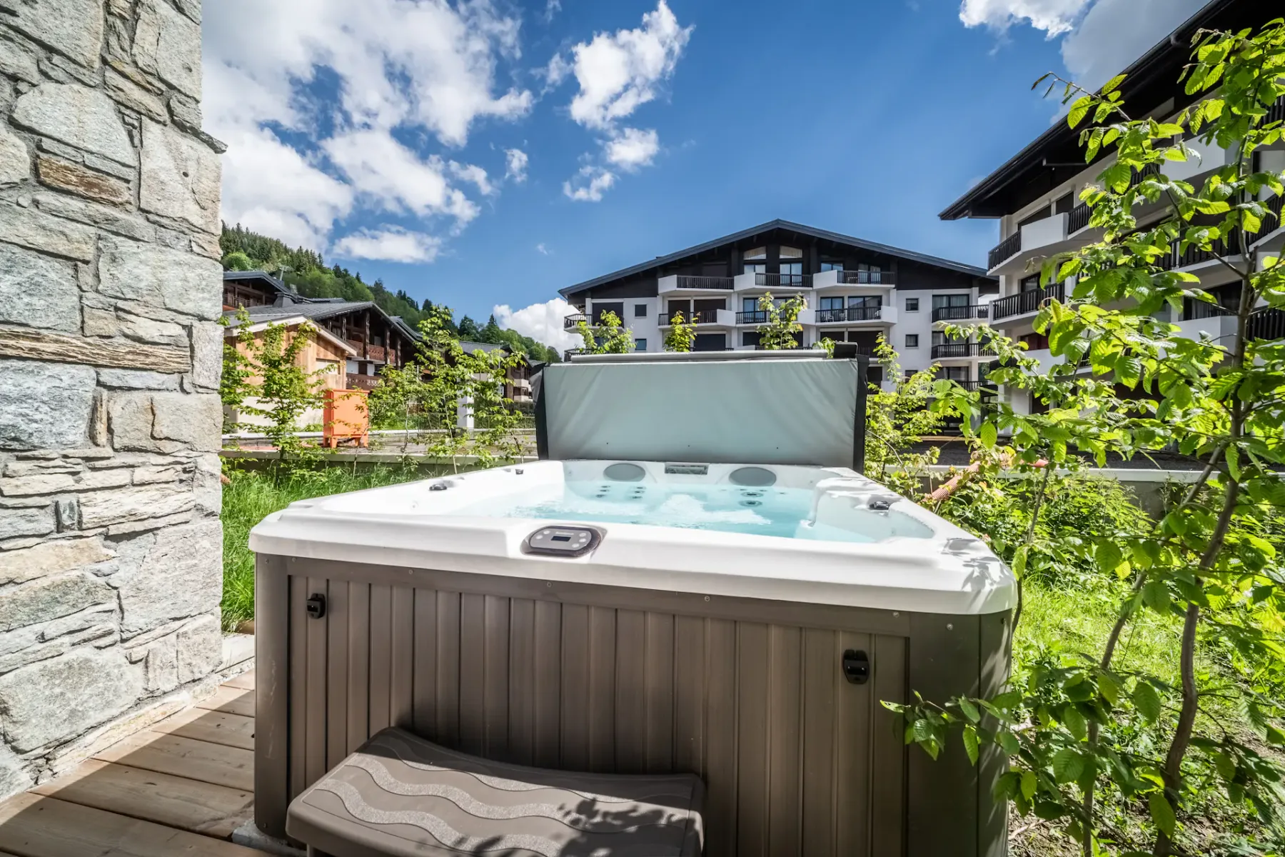 An outdoor hot tub sits beside a stone wall, with greenery and buildings behind it.