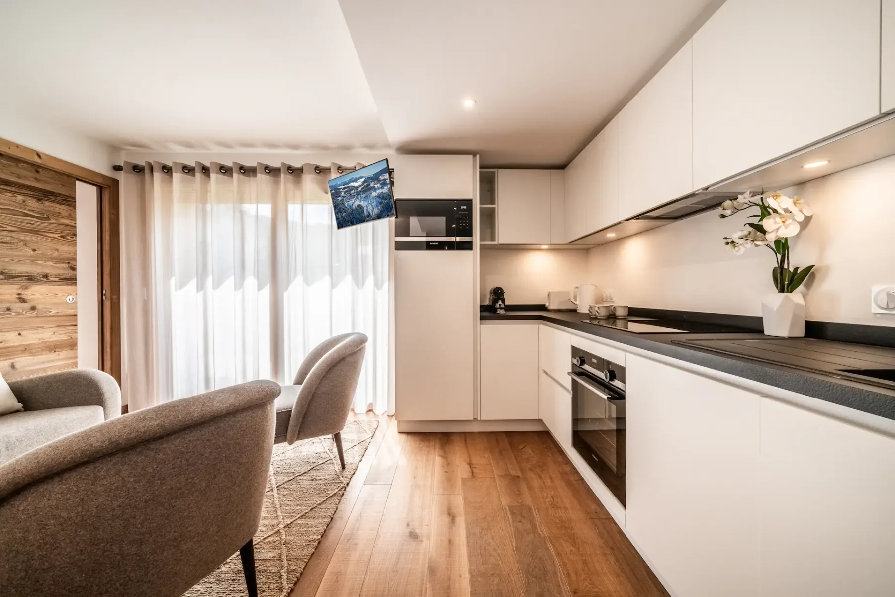 Modern kitchen with white cabinetry, built-in appliances including a microwave, oven, and stove, black countertops, a sink, and wooden flooring. A vase with flowers is placed on the counter.