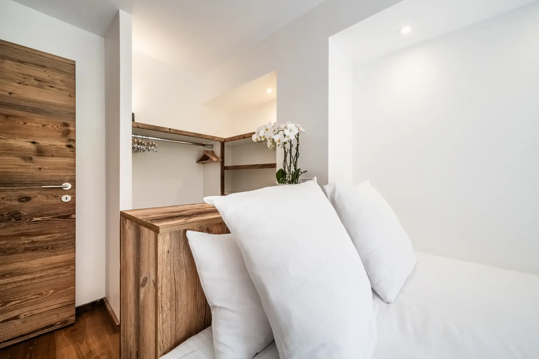 A bedroom with white bedding, wooden cabinetry, and an open hanging area.