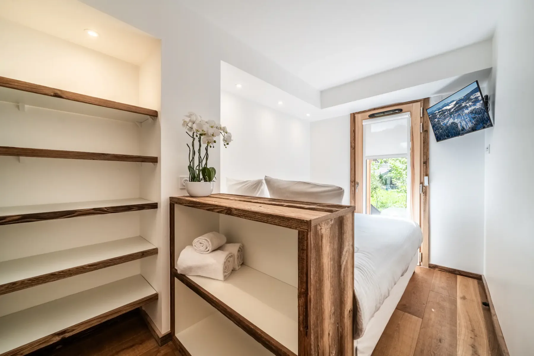 A bright bedroom with white bedding, open wooden shelving, and a window beside the bed.