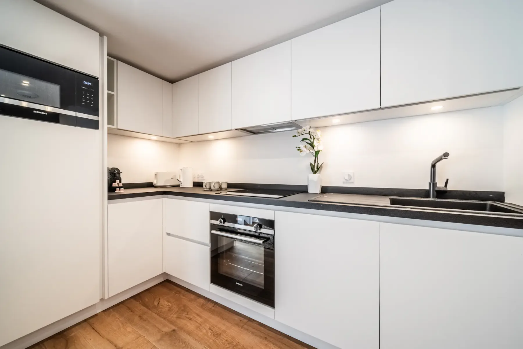 Modern kitchen with white cabinetry, built-in appliances including a microwave, oven, and stove, black countertops, a sink, and wooden flooring. A vase with flowers is placed on the counter.