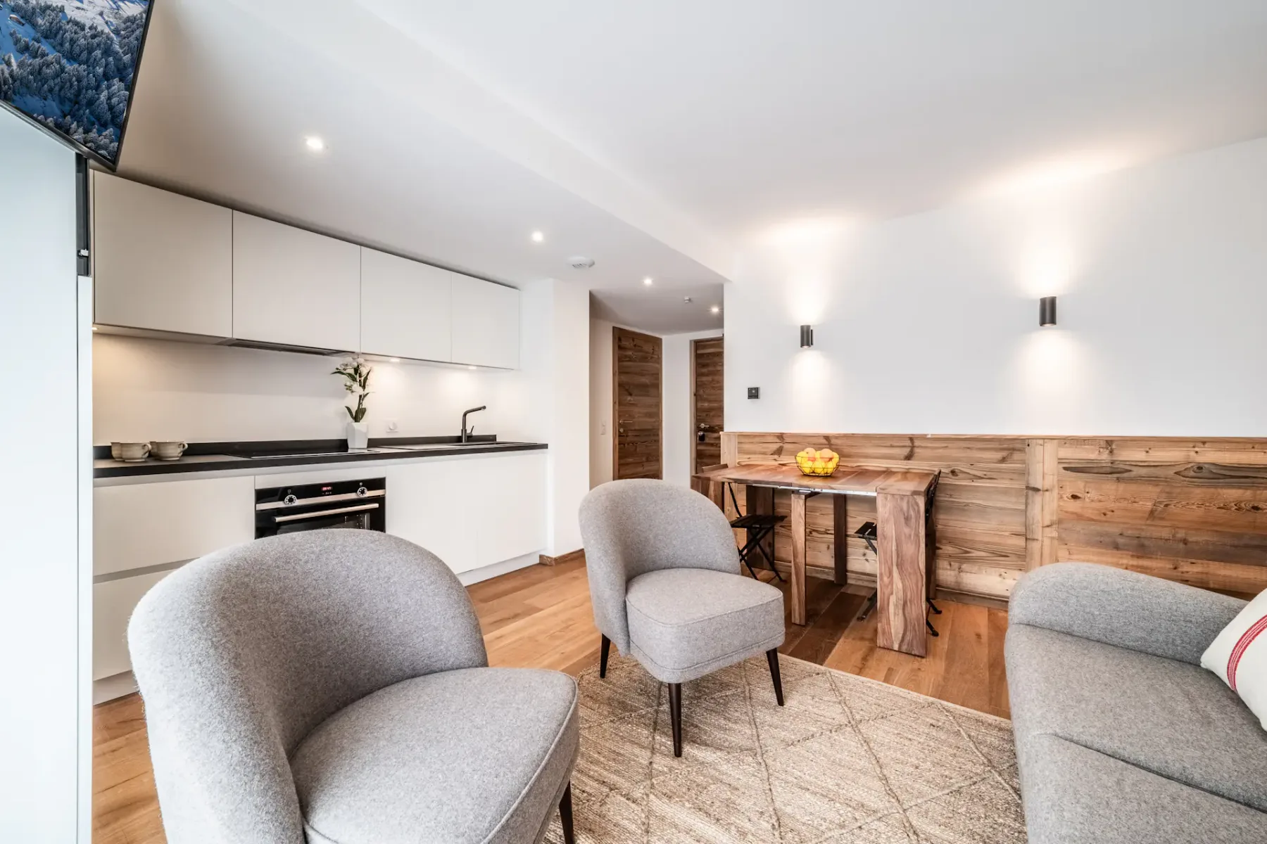 A modern living space featuring a wooden dining table with a fruit bowl, two armchairs, a sofa, a wall-mounted TV, and a kitchenette in a bright room with wooden floors and natural light.