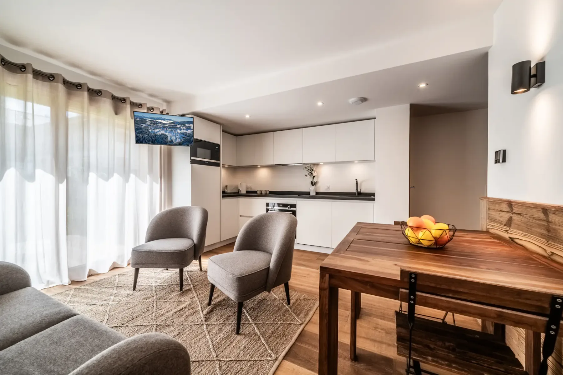 A modern living space featuring a wooden dining table with a fruit bowl, two armchairs, a sofa, a wall-mounted TV, and a kitchenette in a bright room with wooden floors and natural light.