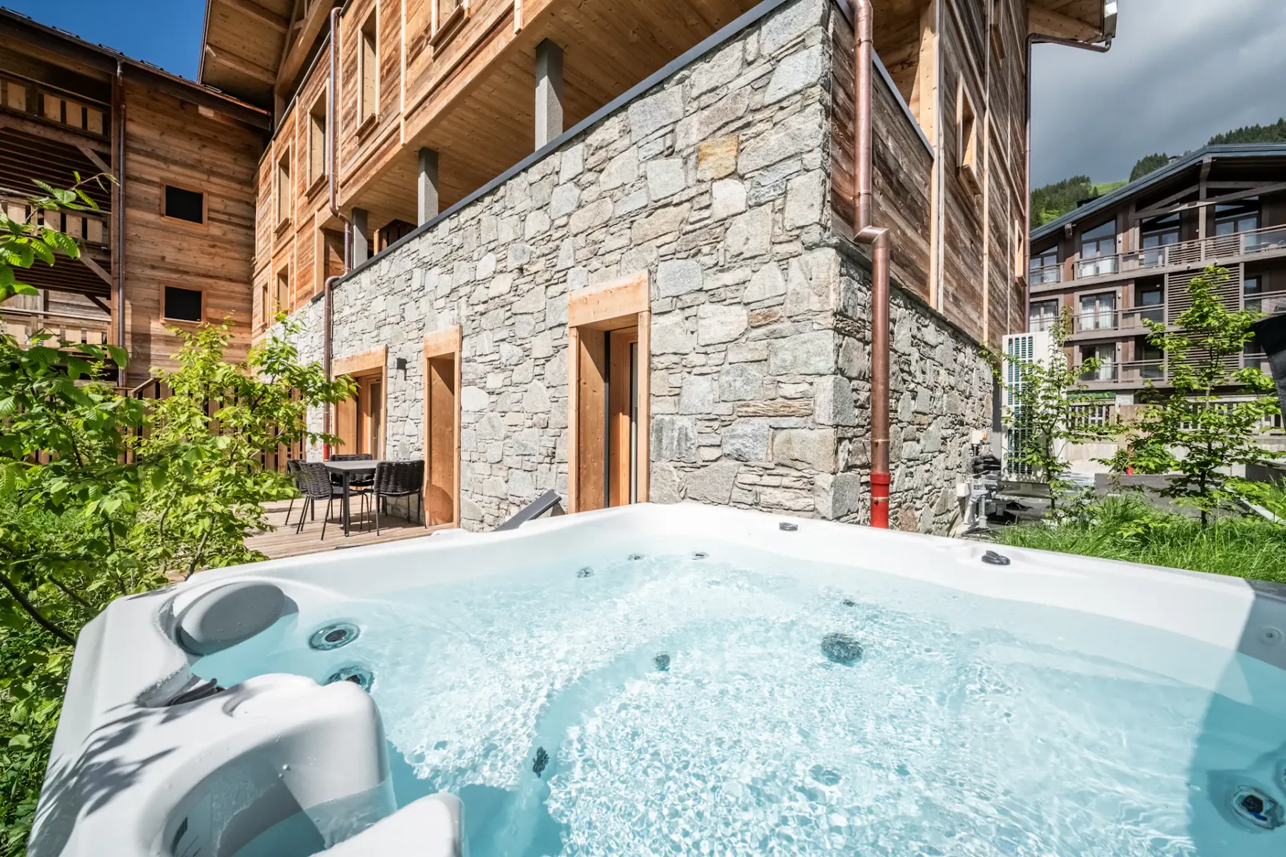 A bubbling outdoor hot tub sits beside a stone wall and timber-clad buildings.