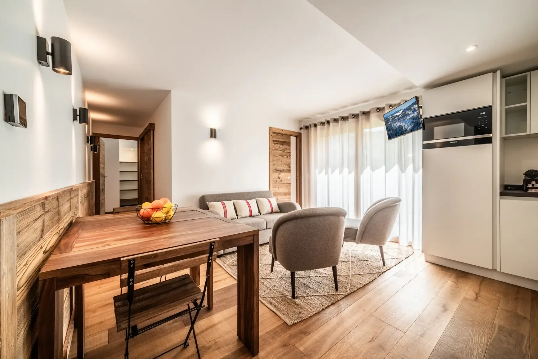 A bright dining and seating area with a wooden table, fruit bowl, and upholstered chairs.