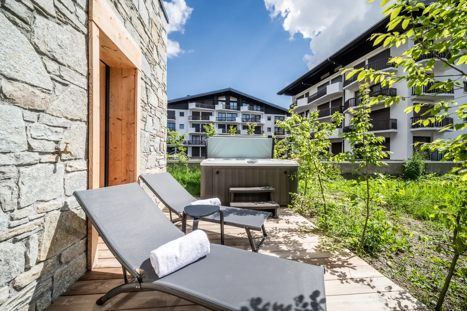 Two gray sun loungers with towels and a small table are placed on a wooden patio beside a hot tub, framed by a stone wall and green plants, with multi-story buildings in the background.