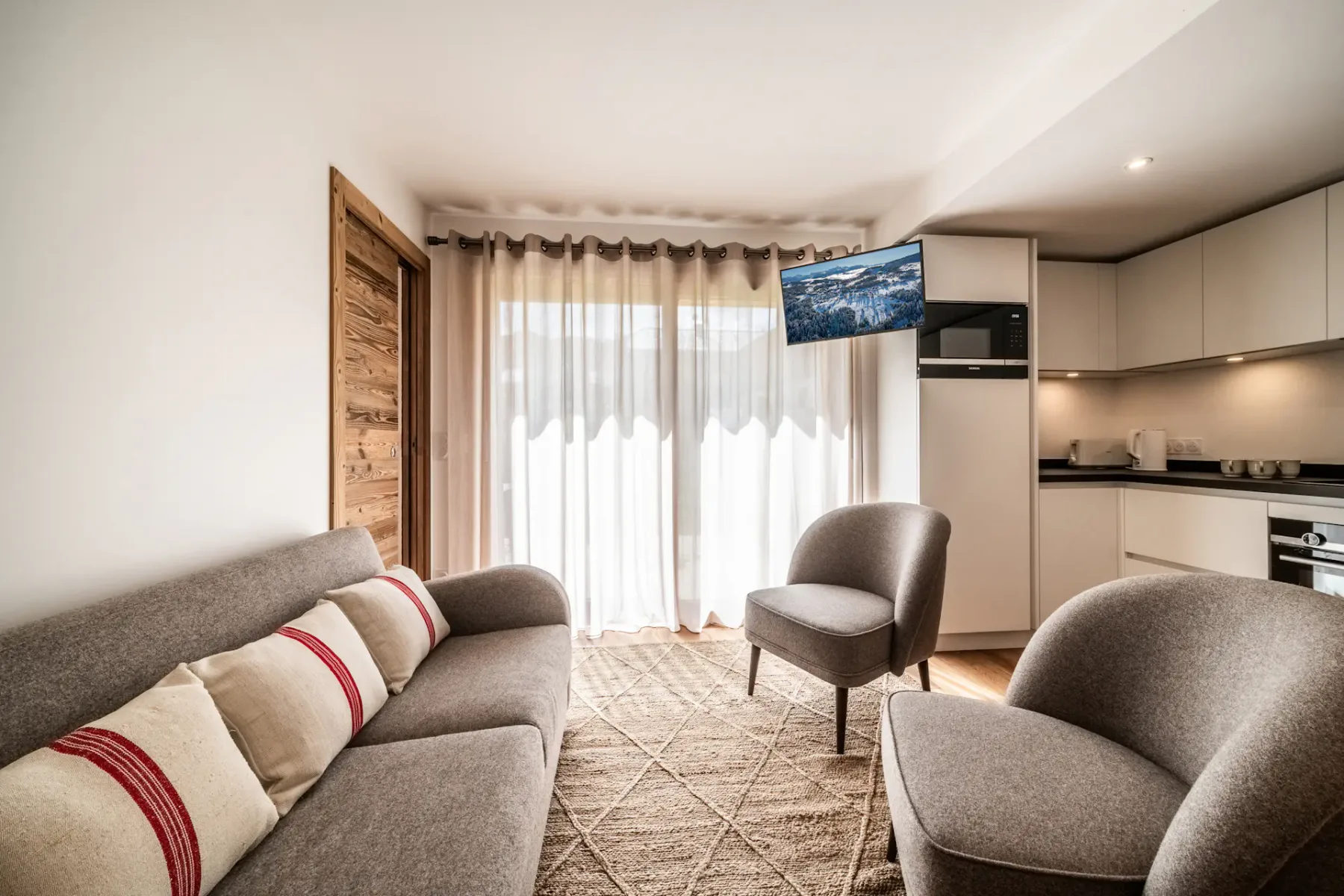 A living area with seating, a television, and a kitchenette beside a curtained window.