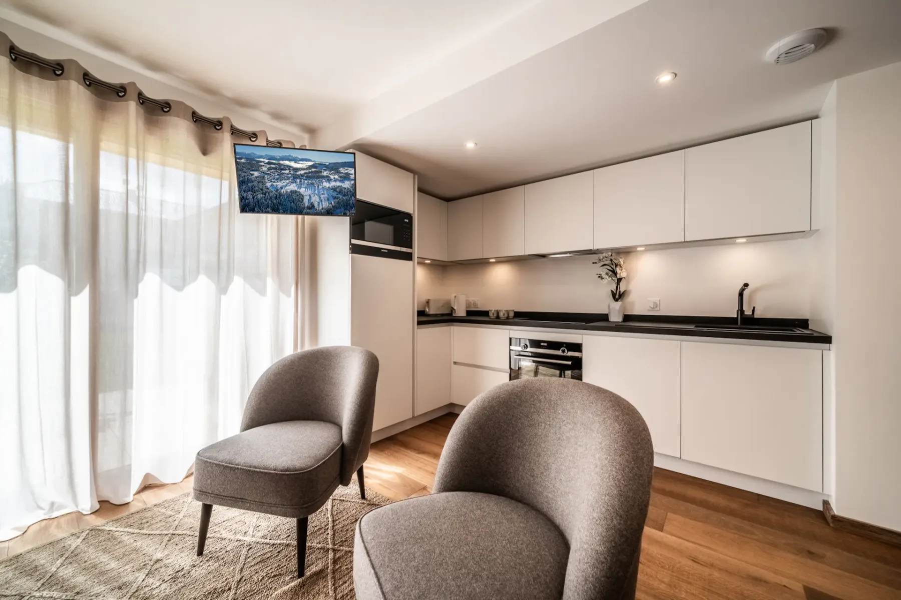 A modern kitchen with white cabinets and dark countertops opens onto a seating area with two chairs.