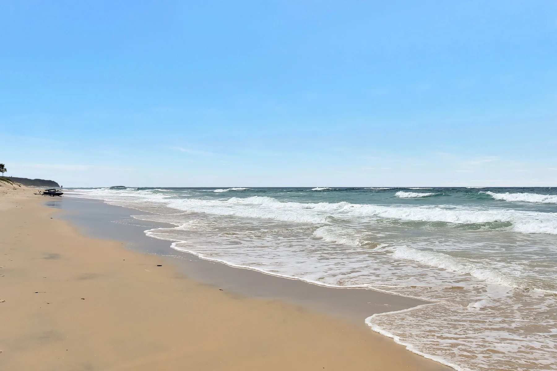 A sandy beach stretches alongside rolling ocean waves under a clear sky.