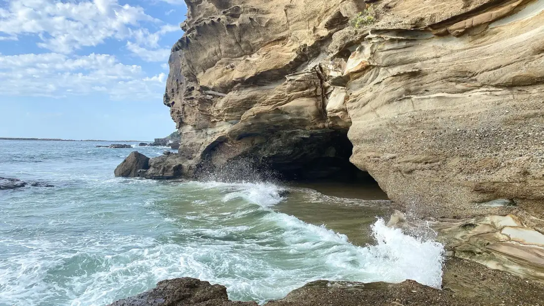 Ocean waves break against a rocky cliff with a dark cave opening.