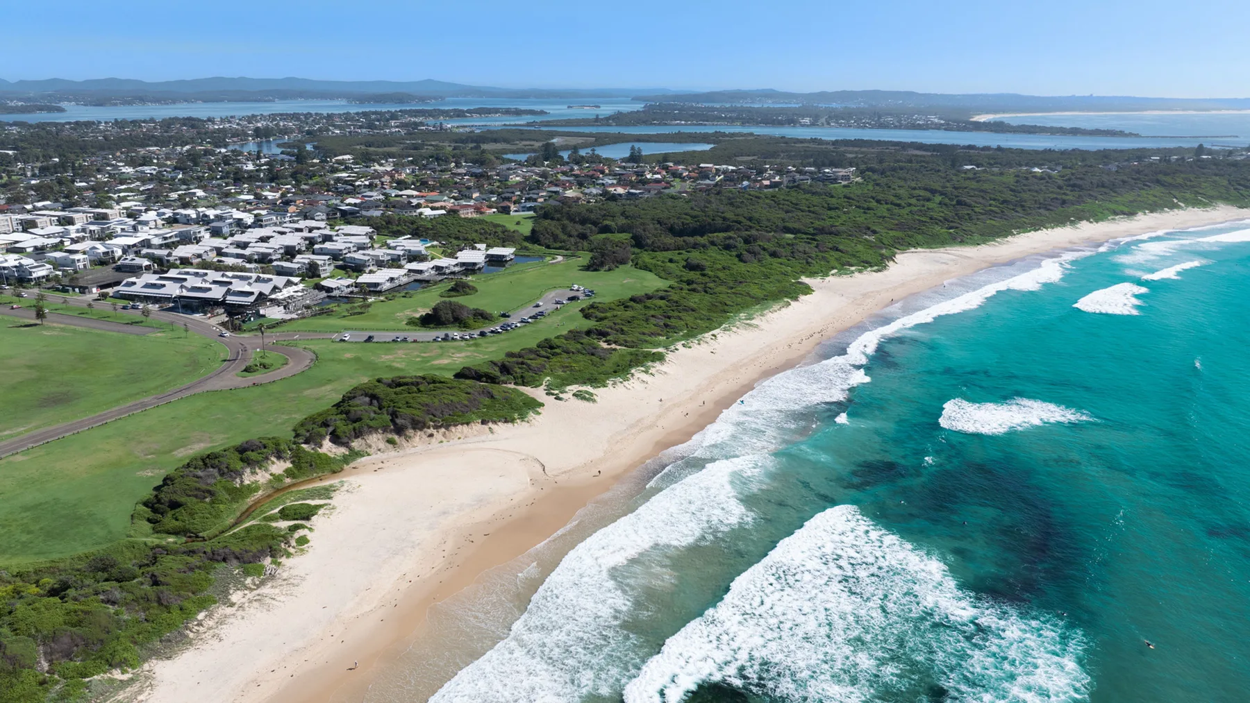 An aerial view shows a sandy beach beside turquoise ocean water and coastal greenery.