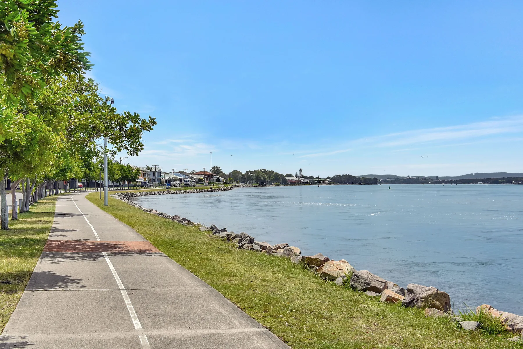 A paved waterfront path runs beside the water and a tree-lined grassy bank.