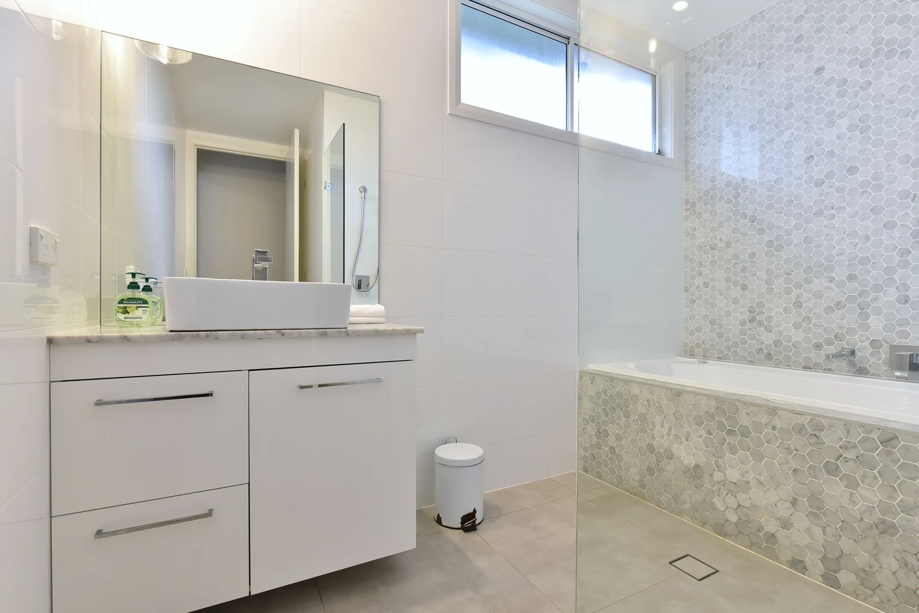 A bright bathroom with a vanity, mirror, and tiled bathtub surround.