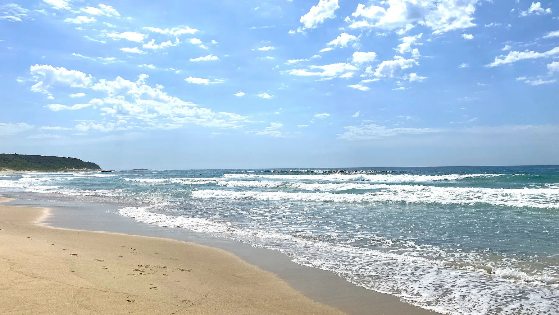 A sandy beach meets the surf beneath a partly cloudy sky.