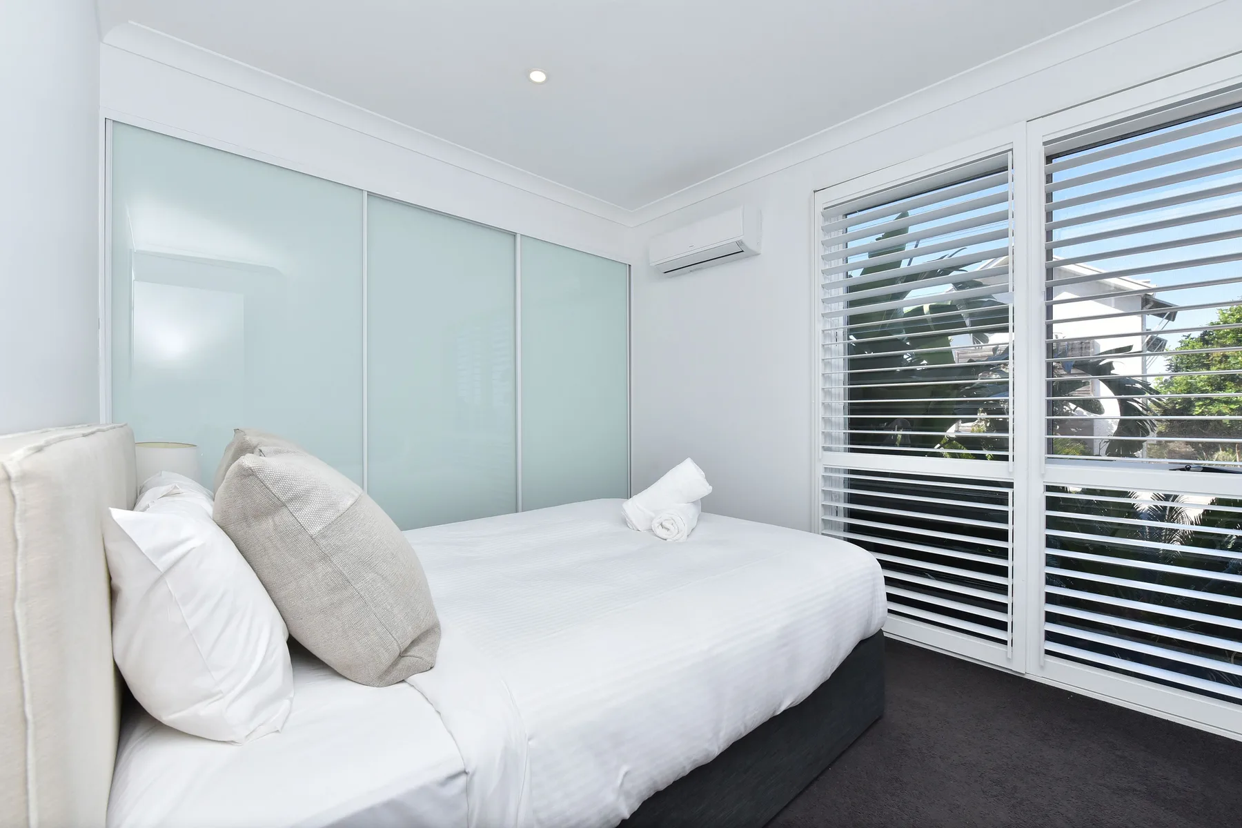 A bright bedroom with a bed beside large windows fitted with white shutters.