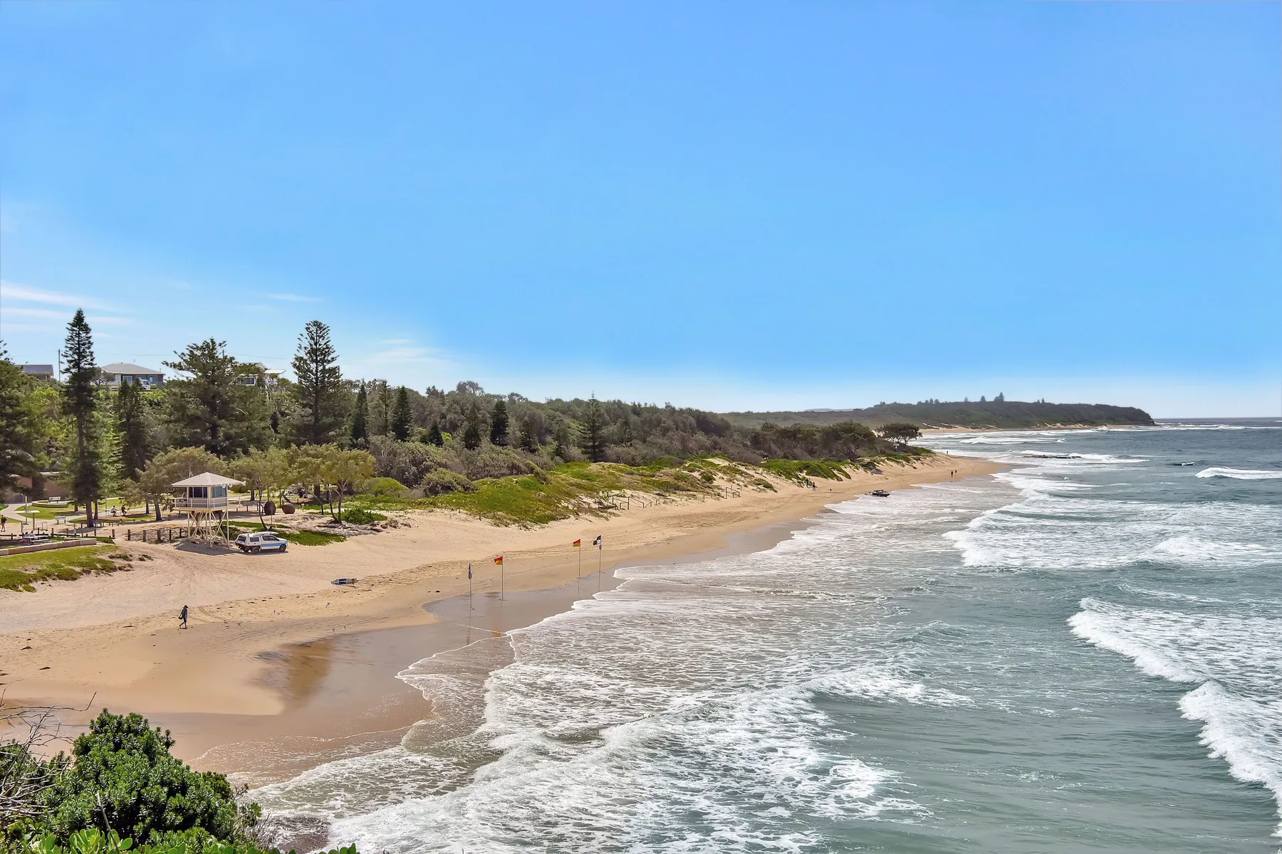 A sandy beach curves along the shoreline beside trees and breaking waves.