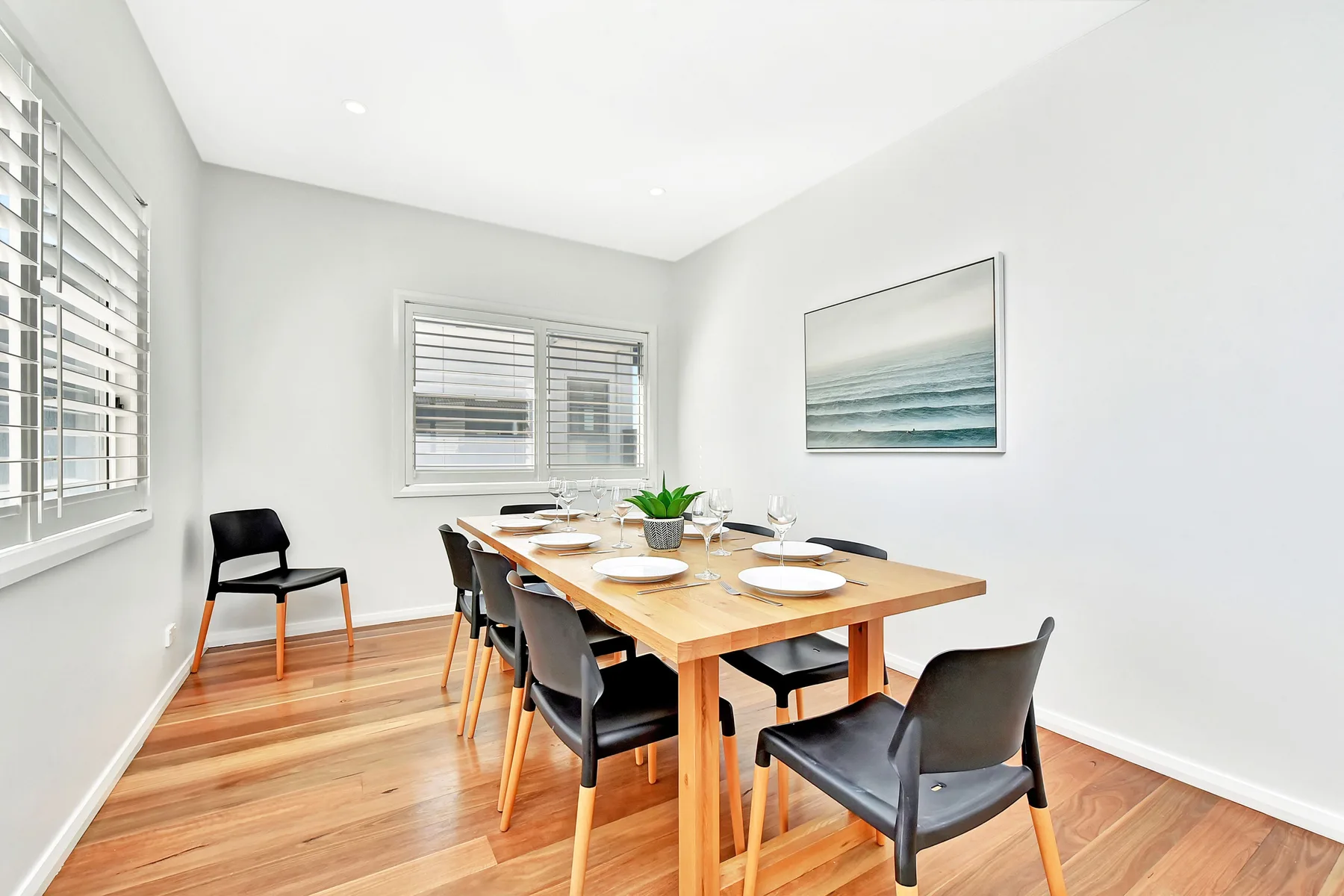 Dining area with a long wooden table and chairs beneath a coastal painting.