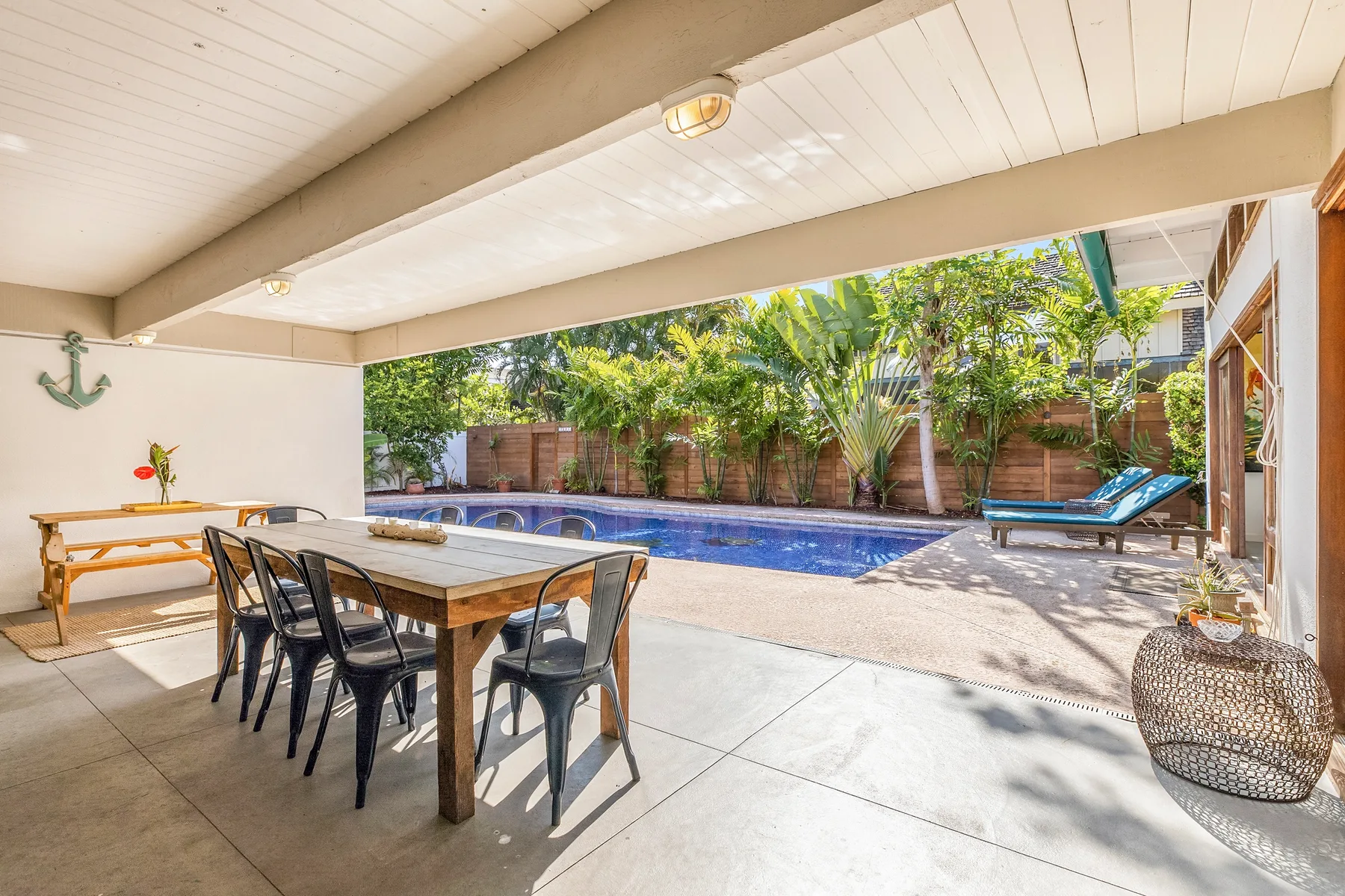 Un patio cubierto con asientos para comer da a una piscina y un jardín.