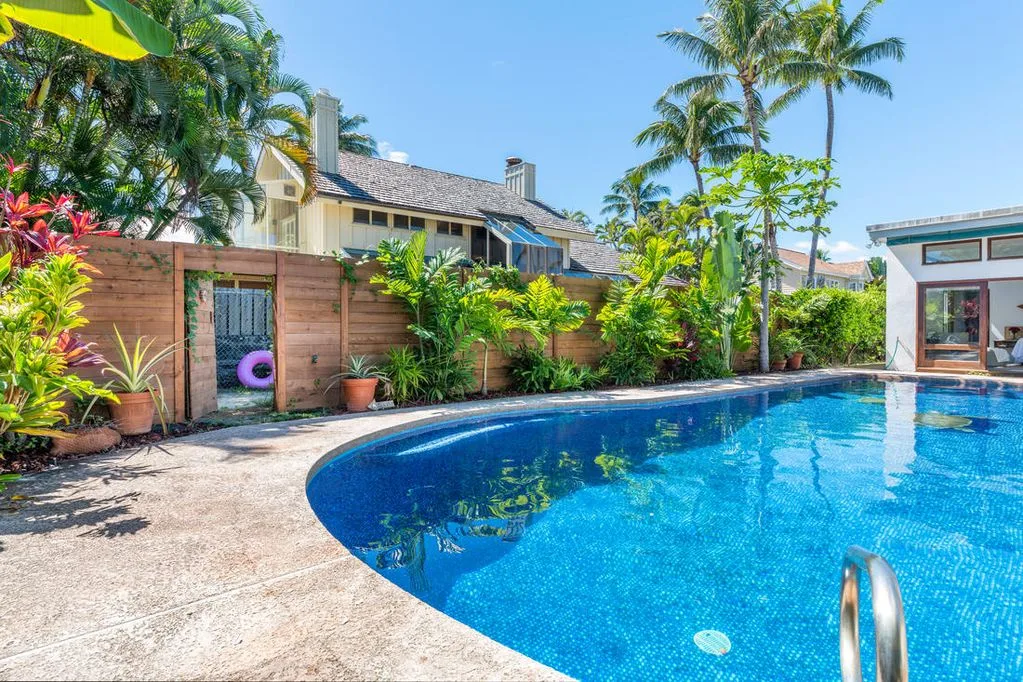Una piscina al aire libre junto a una terraza pavimentada, plantas tropicales y una cerca de madera.