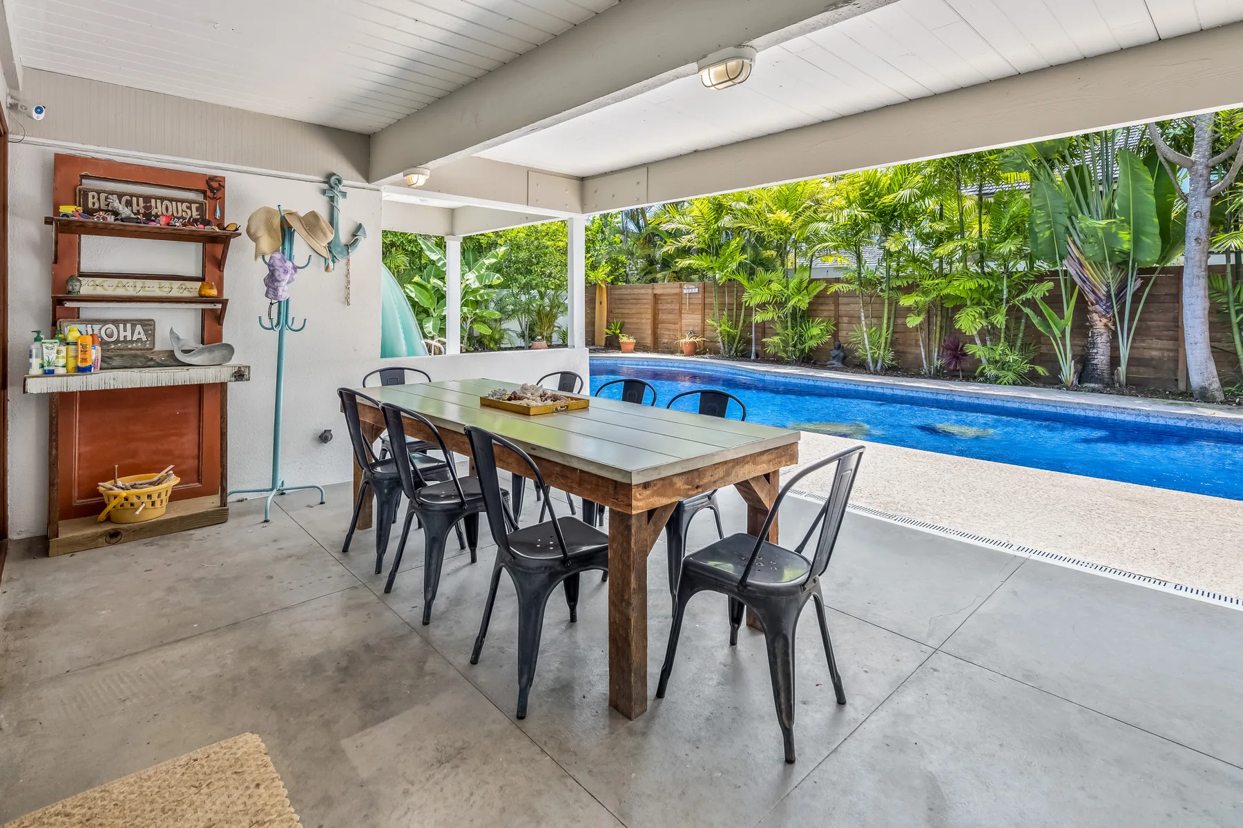 A covered patio has a long dining table beside a swimming pool.