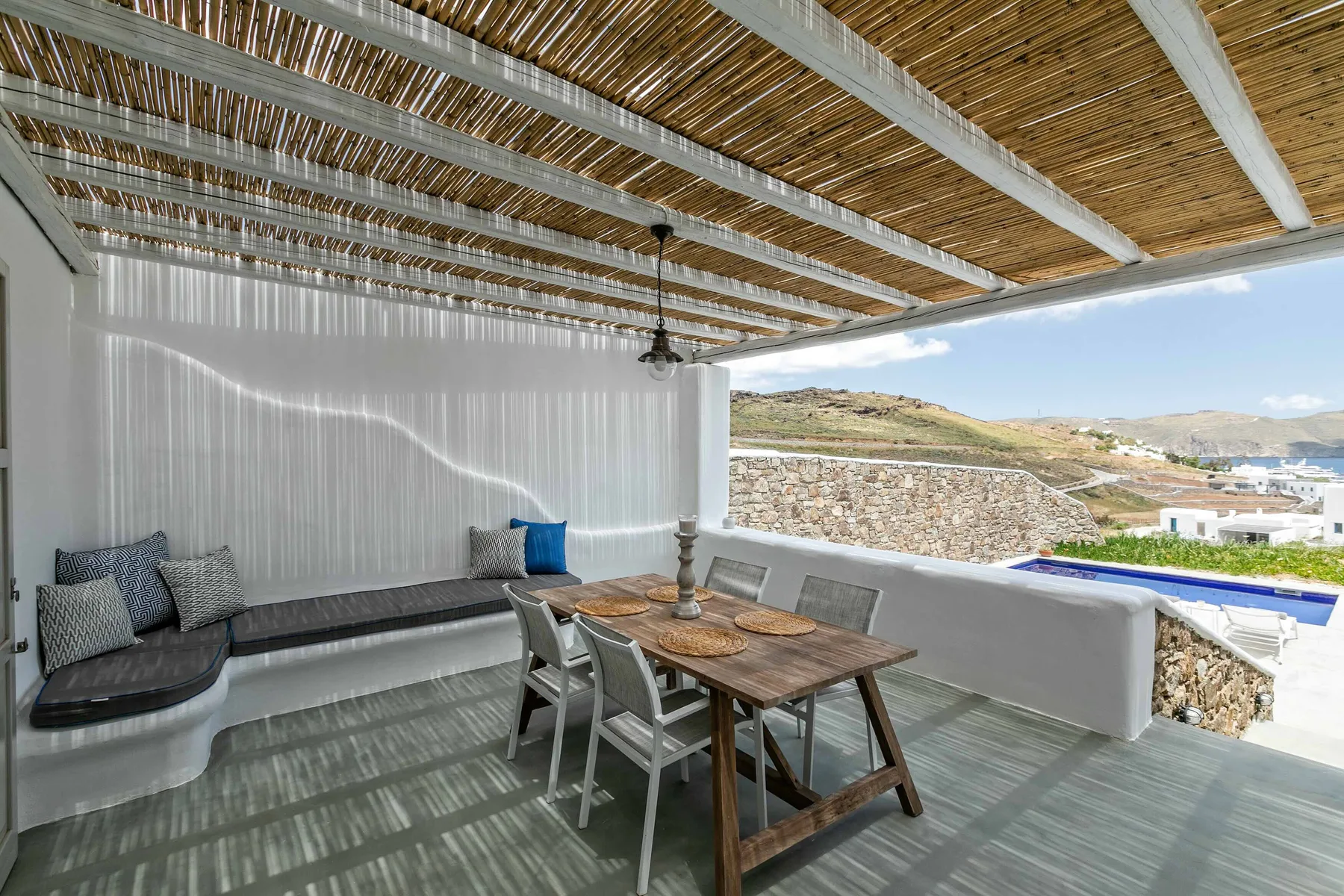 Una terraza cubierta tiene una mesa de comedor y asientos con vistas a un paisaje costero.