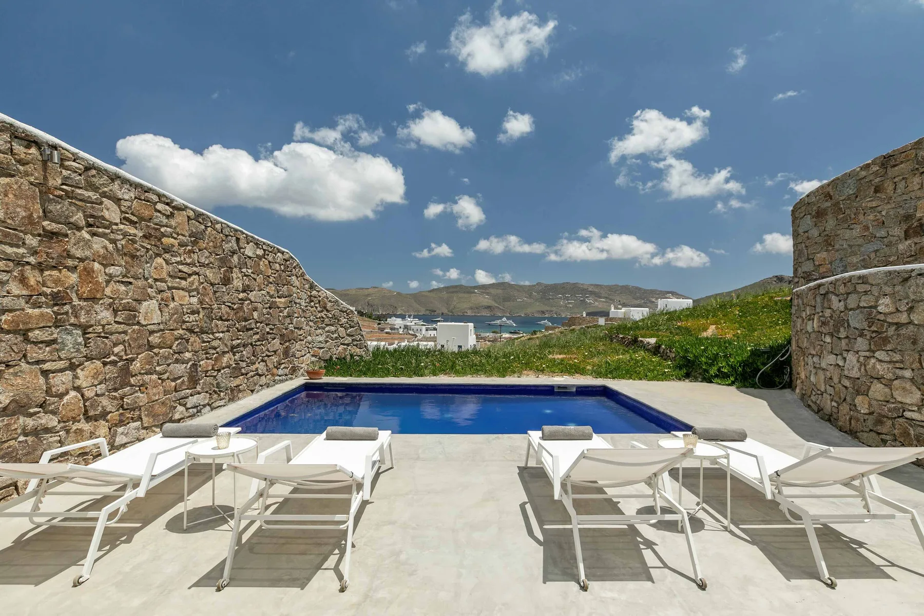 Una piscina al aire libre rodeada de muros de piedra, con asientos y vistas al mar a lo lejos.