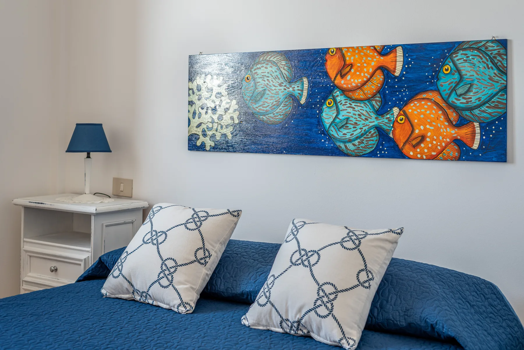 Un dormitorio con ropa de cama azul, cojines estampados y un cuadro colorido de peces sobre la cama.