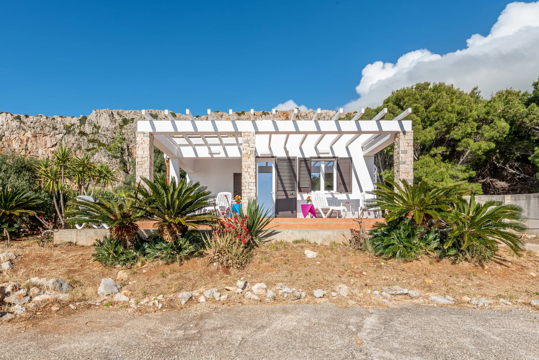 Una casa baja de piedra con una pérgola blanca está rodeada de plantas desérticas.