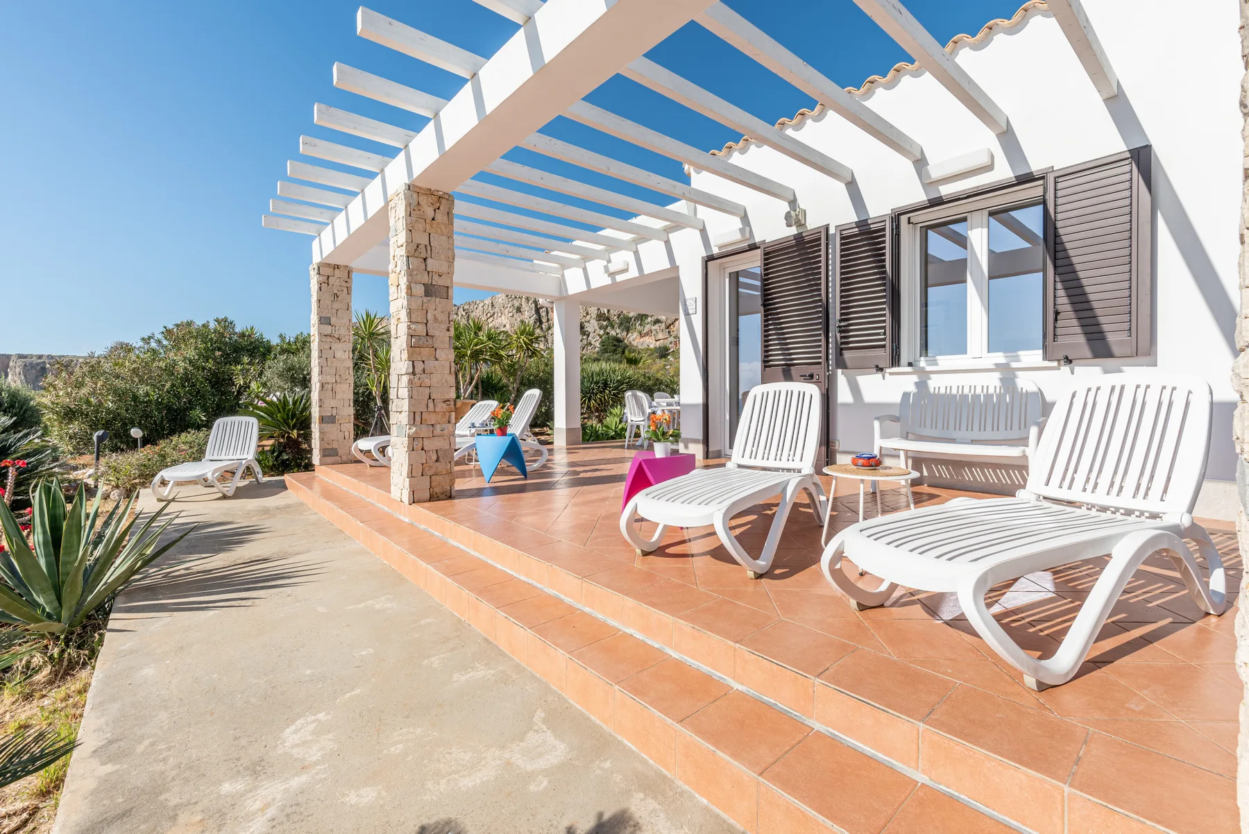 A sunny outdoor patio with white lounge seating beneath a pergola.