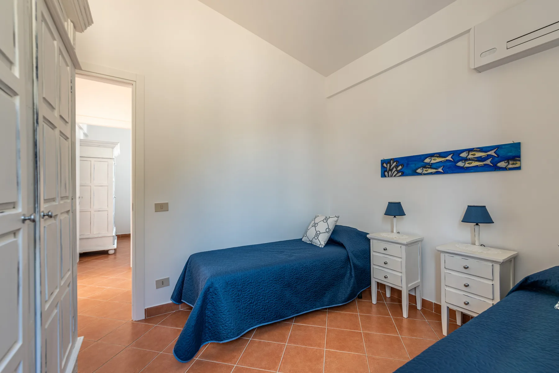 Un dormitorio con camas cubiertas de azul, mesitas de noche y suelo de baldosas.