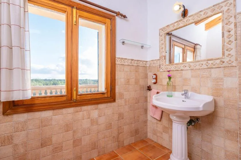 Un baño alicatado con lavabo de pedestal, espejo y ventana con marco de madera.