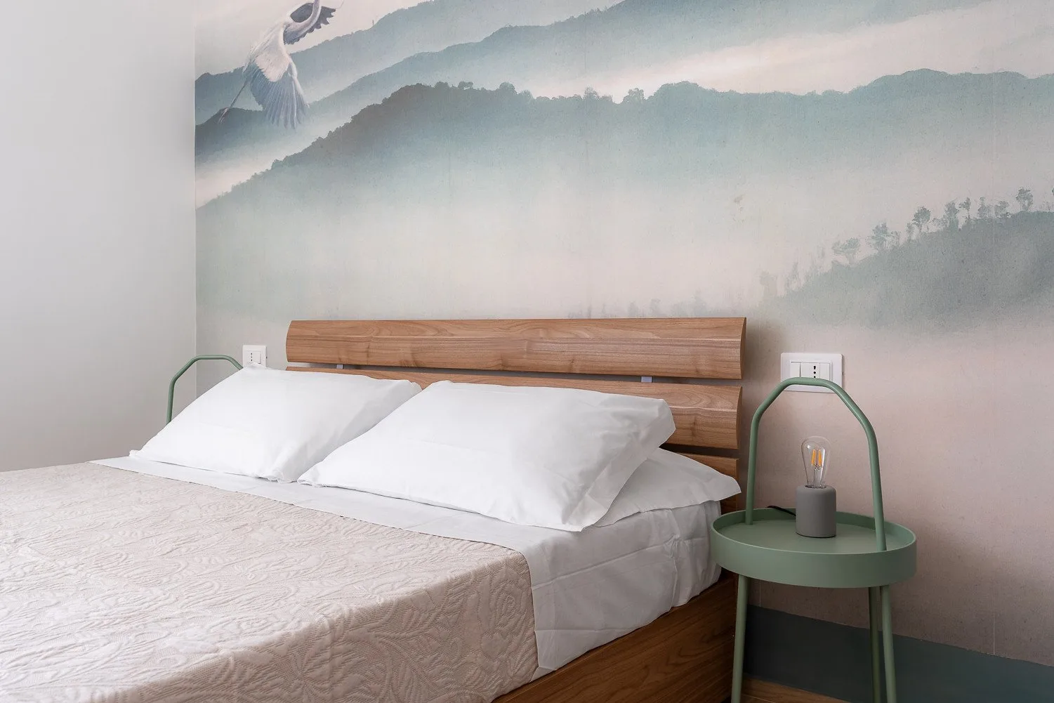 Un dormitorio con ropa de cama blanca y un cabecero de madera bajo un mural de montañas brumosas.