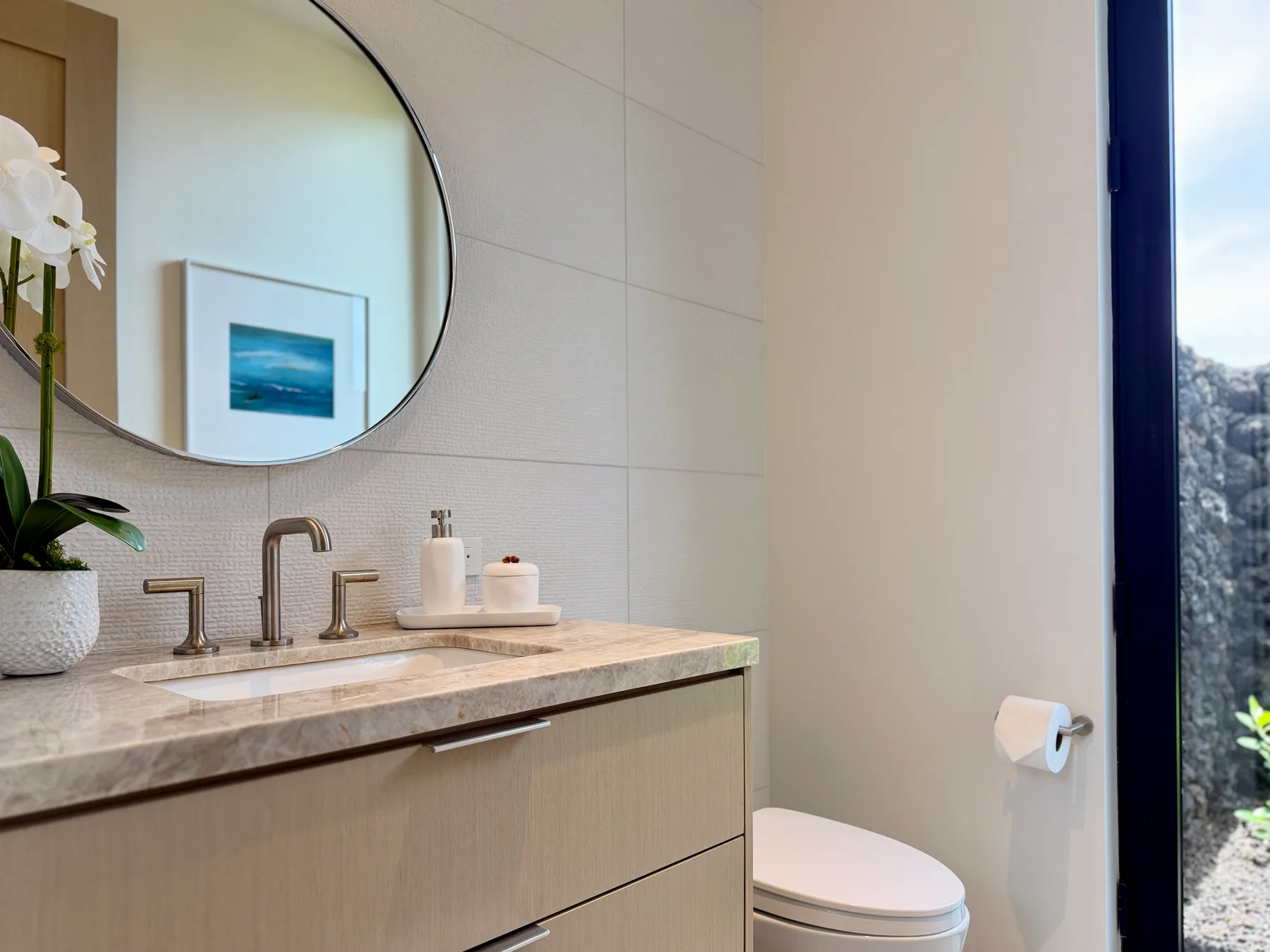 Un baño luminoso con un lavabo, un espejo redondo grande y un inodoro junto a la ventana.