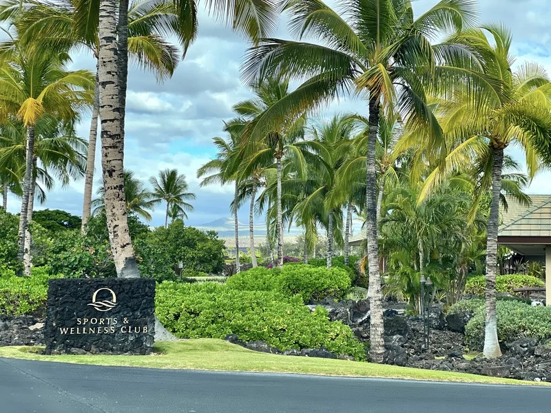 Una entrada tropical ajardinada muestra palmeras, vegetación y un letrero de piedra.