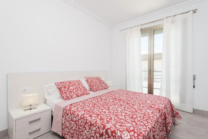 A bedroom with a double bed covered in red floral bedding beside a curtained window.