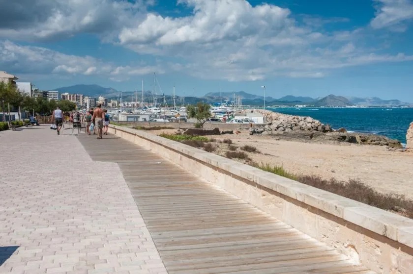 A seaside promenade runs beside a beach, with boats and mountains visible across the bay.