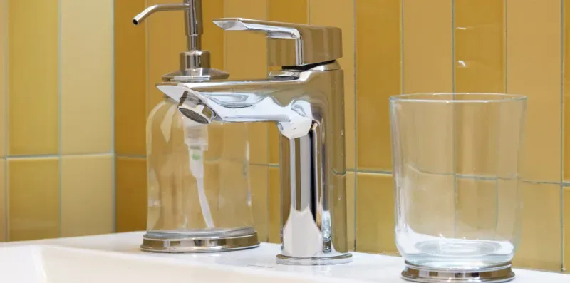 Un grifo de baño y un vaso están frente a paredes de azulejos amarillos.