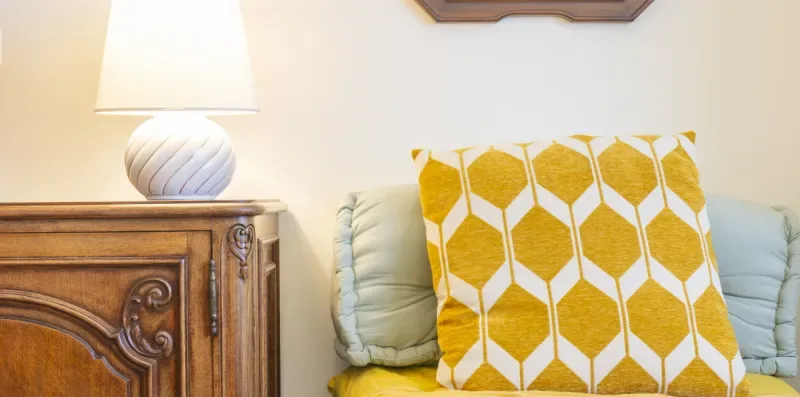 Una zona de asientos con un cojín amarillo estampado junto a un mueble de madera y una lámpara.