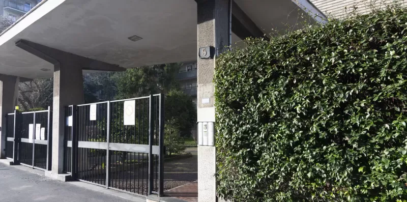 Una entrada con reja está bajo una estructura cubierta junto a un seto alto.