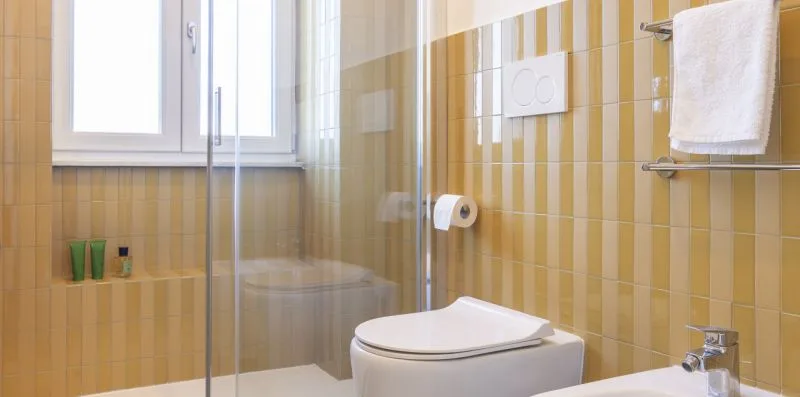 Un baño con azulejos, inodoro, ducha de vidrio y una ventana luminosa.