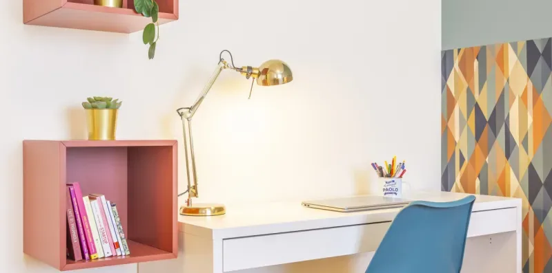 Un escritorio con una silla azul, una lámpara dorada y estantes para libros junto a una pared blanca.