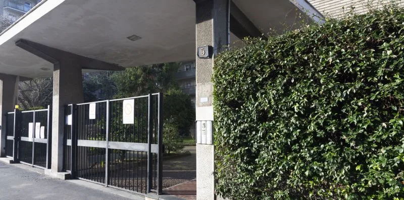 Una entrada con reja está bajo un voladizo de hormigón, junto a un seto alto.