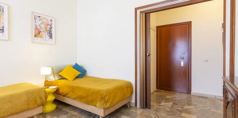 Un dormitorio con una cama color mostaza, una mesita amarilla y cuadros en la pared.