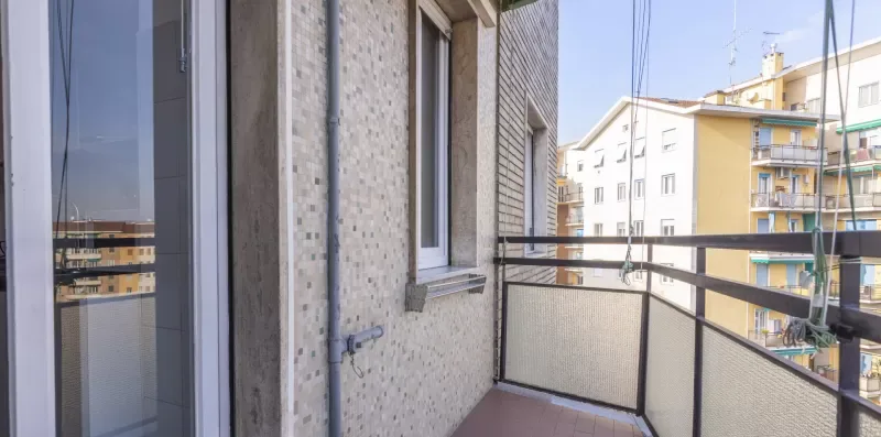 Un balcón estrecho con barandilla metálica da a edificios de apartamentos cercanos.