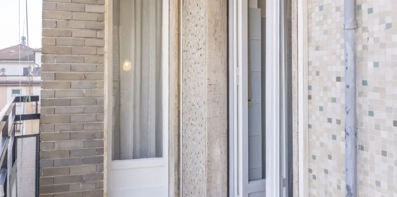 Un balcón alicatado con puertas corredizas de vidrio y edificios vecinos visibles afuera.