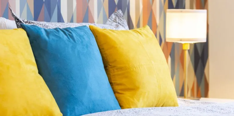 Una cama con almohadas amarillas y azules destaca frente a una pared de colores y estampados.