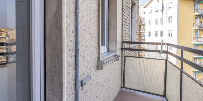 Un balcón estrecho con barandilla, paredes de baldosas y una ventana frente a edificios cercanos.
