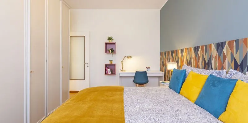 Un dormitorio con cojines coloridos sobre la cama y un escritorio junto a la pared del fondo.